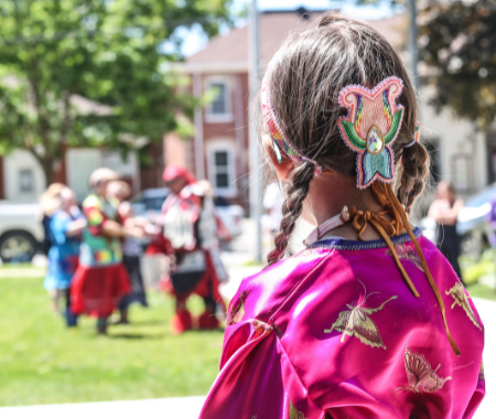 A brown haired girl with her hair in two braids. The image shows the back of her head. She is wearing a colourful beaded barrette and a purple shirt. The background of the image shows blurred people.